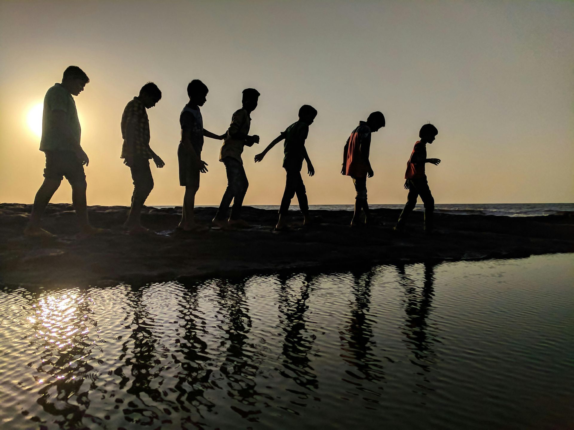 Silhuetter af mennesker, der går langs vandet ved solnedgang; refleksion synlig.