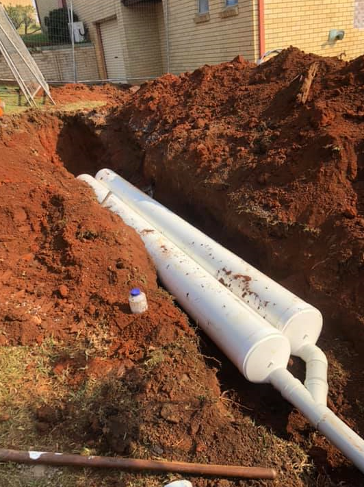 A trench in reddish soil contains two white PVC pipes connected by a Y-junction, with a house visible in the background — B&A Plumbing and Gas In Wauchope, NSW