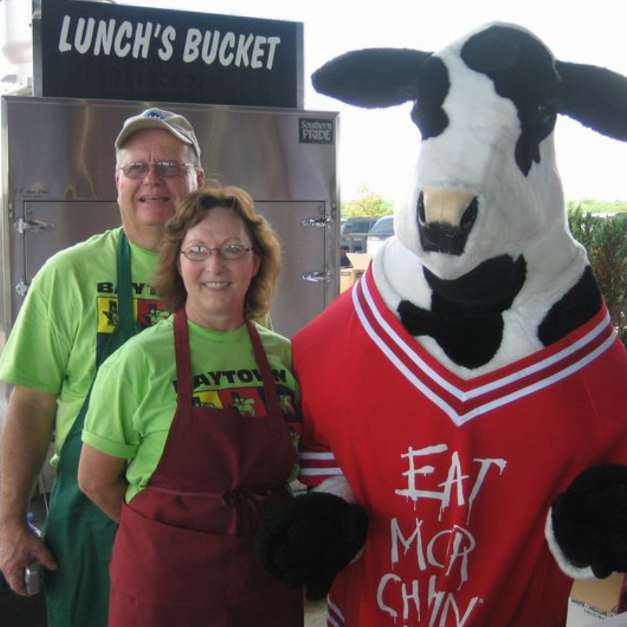 Two people and a cow mascot in red jersey at a lunch stand labeled “Lunch’s Bucket”
