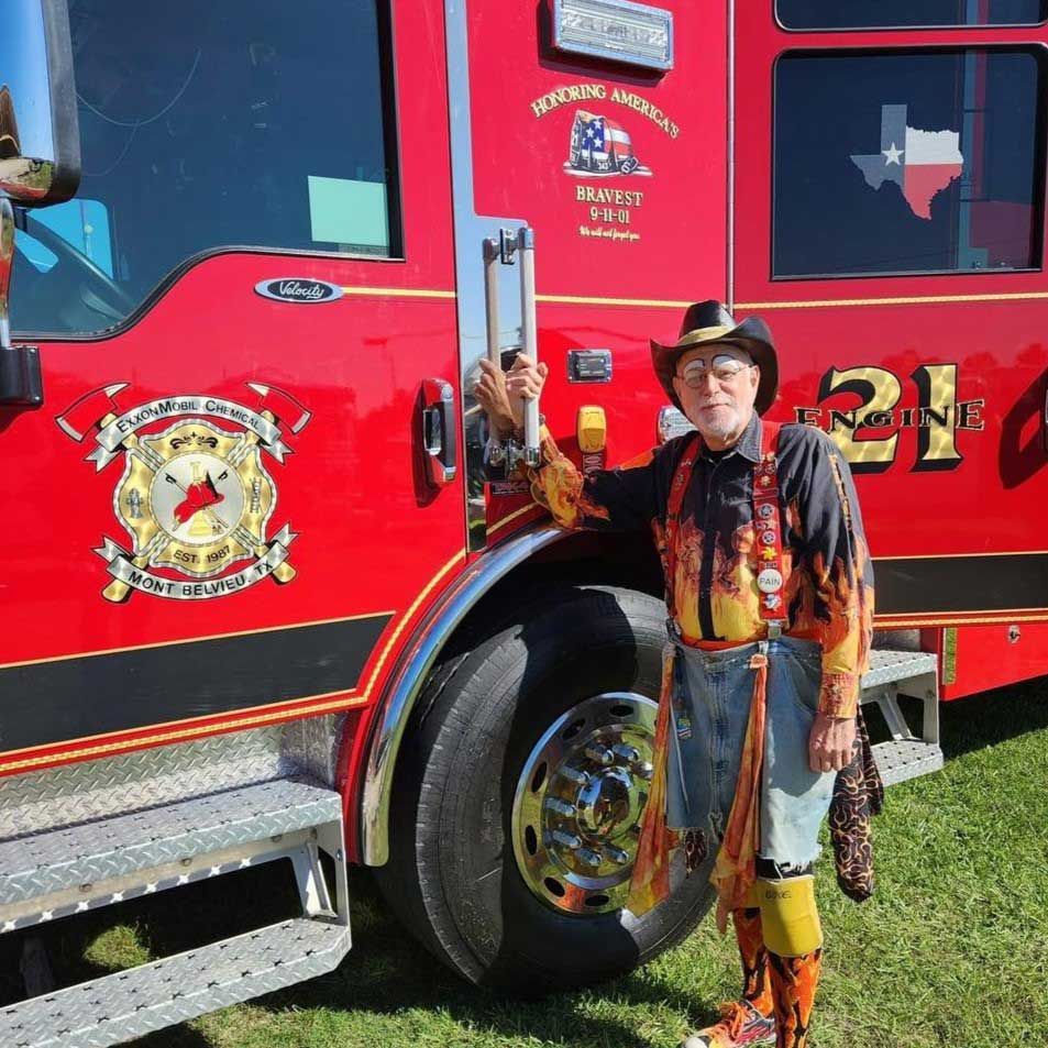 A man in clown makeup and a cowboy hat and fiery-patterned clothing stands next to a red fire engine for ExxonMobil Chemical Mont Belvieu during the Baytown Special Rodeo.