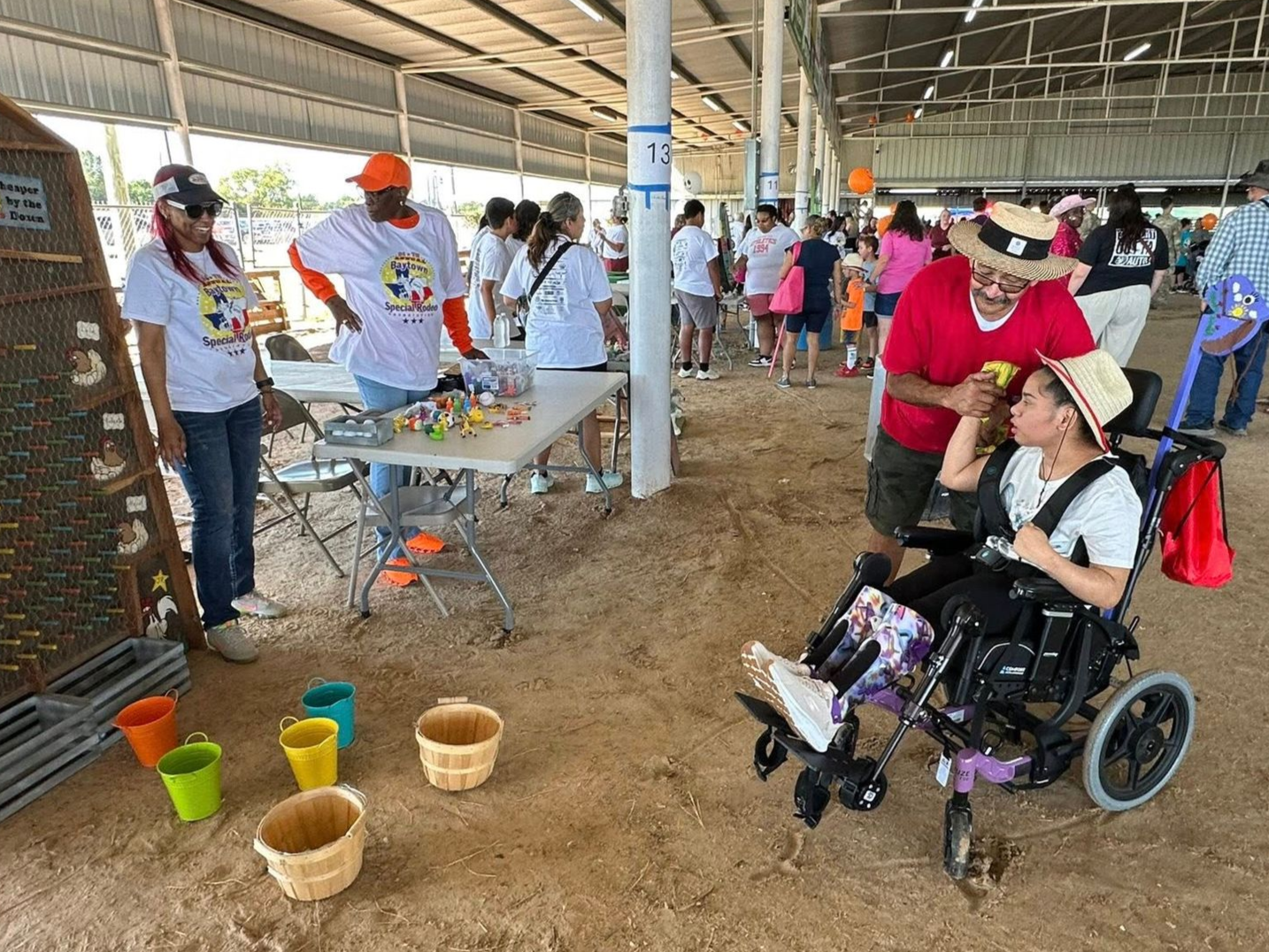 A person in a wheelchair is being helped by another person at an outdoor event with various stalls and people in the background during the Baytown Special Rodeo.