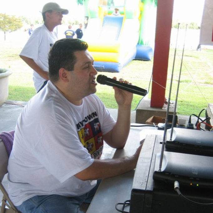 Man speaking into a microphone at an outdoor event booth, with audio equipment and a bouncy castle in the background