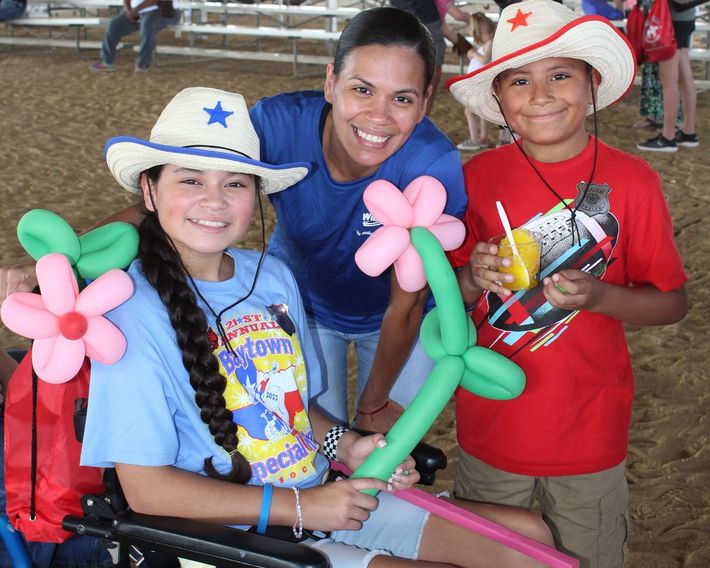 Two children holding balloon flowers and a drink stand with a smiling woman at the Baytown Special Rodeo. The children are wearing cowboy hats.