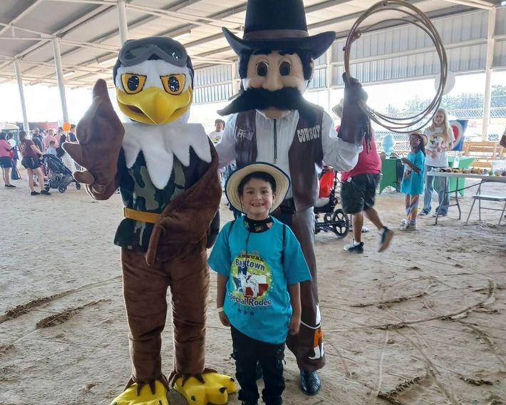 A child in a cowboy hat and blue t-shirt stands between two mascots, an eagle and a cowboy holding a lasso, at the Baytown Special Rodeo.