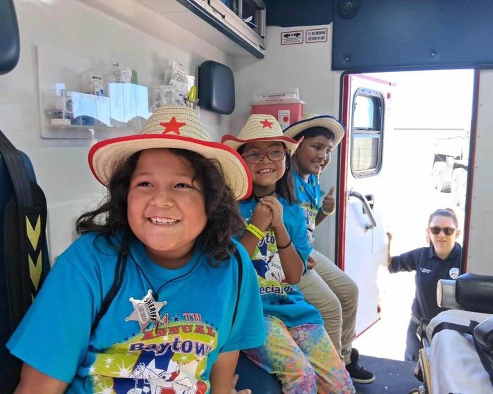 Three smiling children wearing cowboy hats and blue shirts sit inside an ambulance, with a medical professional standing in the doorway during the Baytown Special Rodeo.