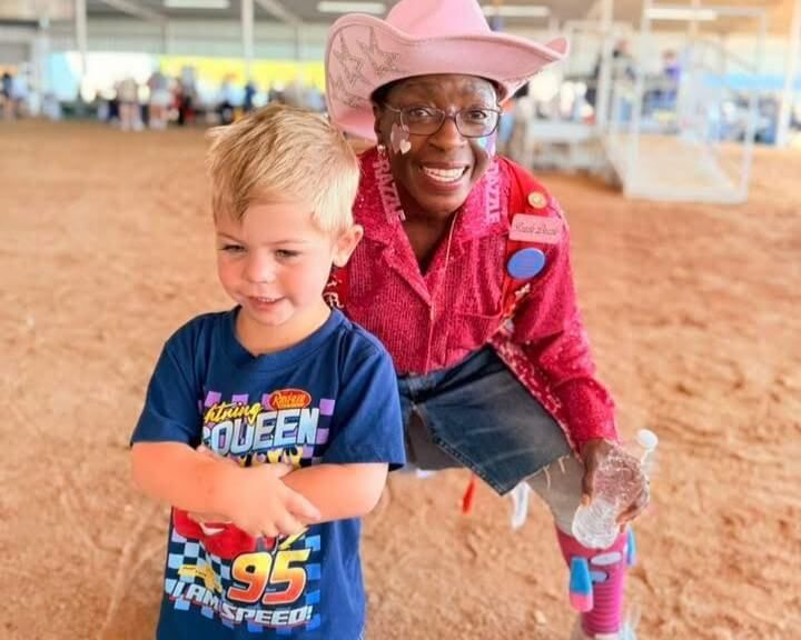 A child in a blue shirt stands next to a woman dressed as a clown in a pink sequined shirt and pink cowboy hat, both smiling at the Baytown Special Rodeo.