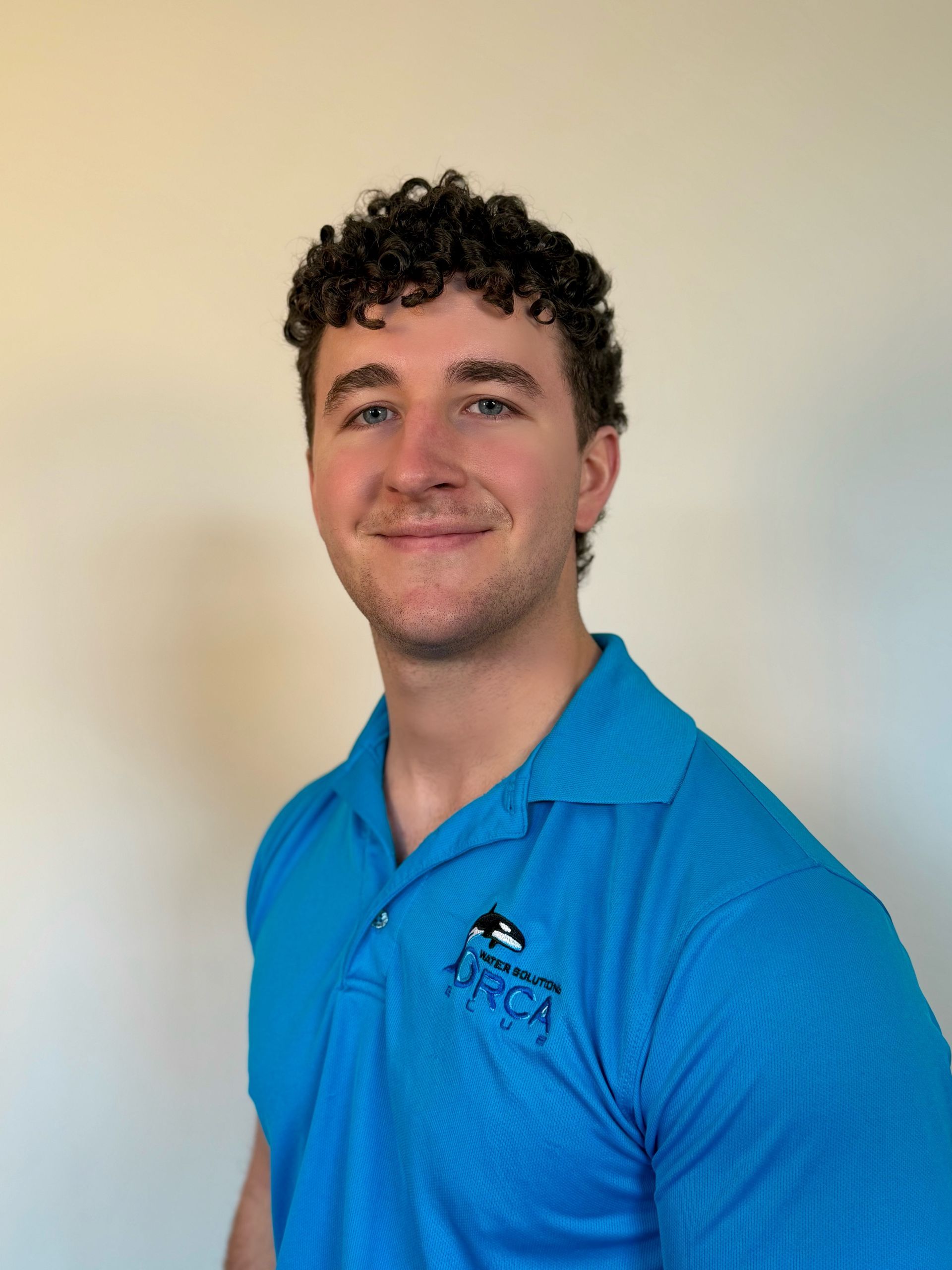 Smiling person in a blue polo shirt standing against a plain beige wall