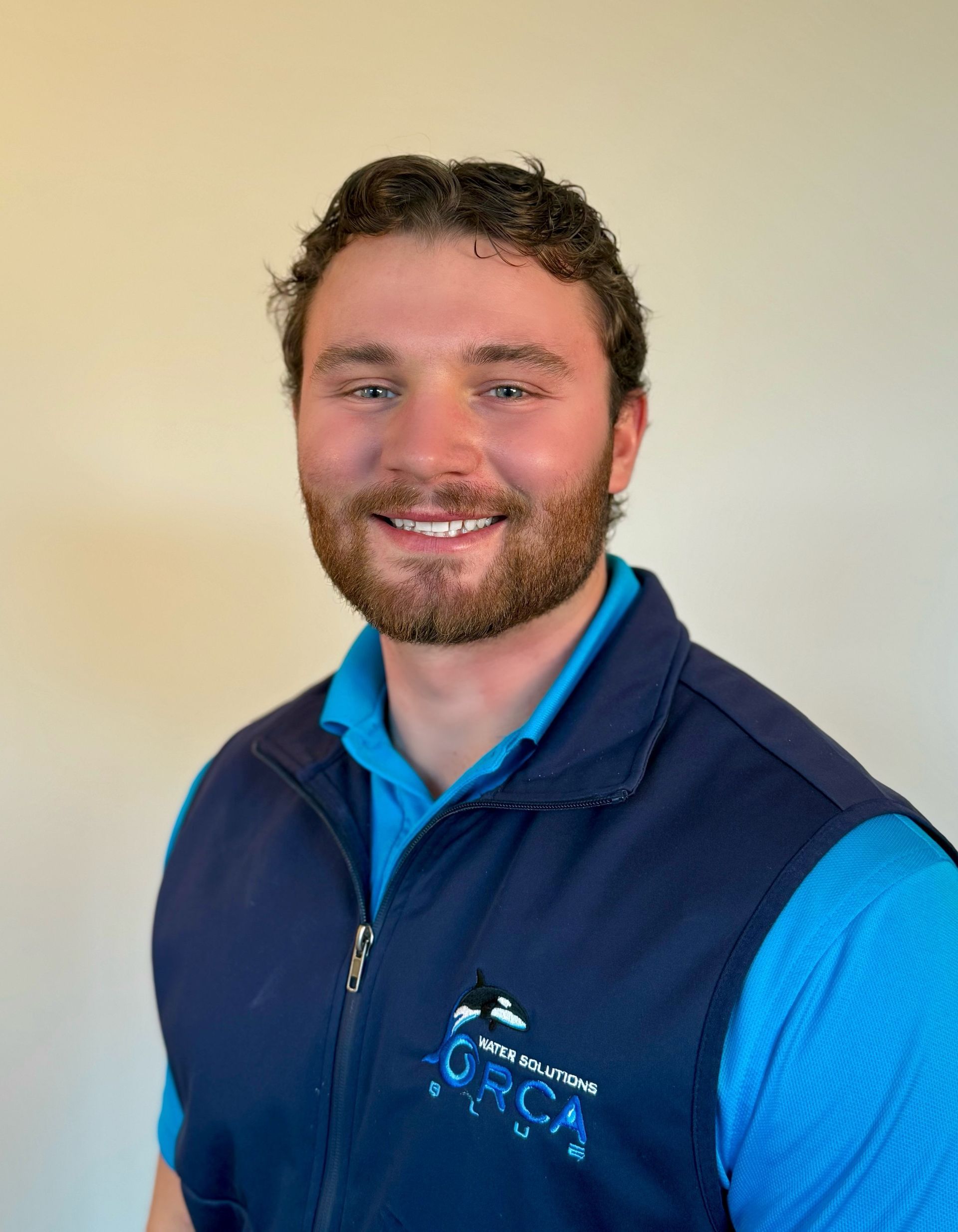 Smiling person in a blue shirt and navy vest against a plain beige background
