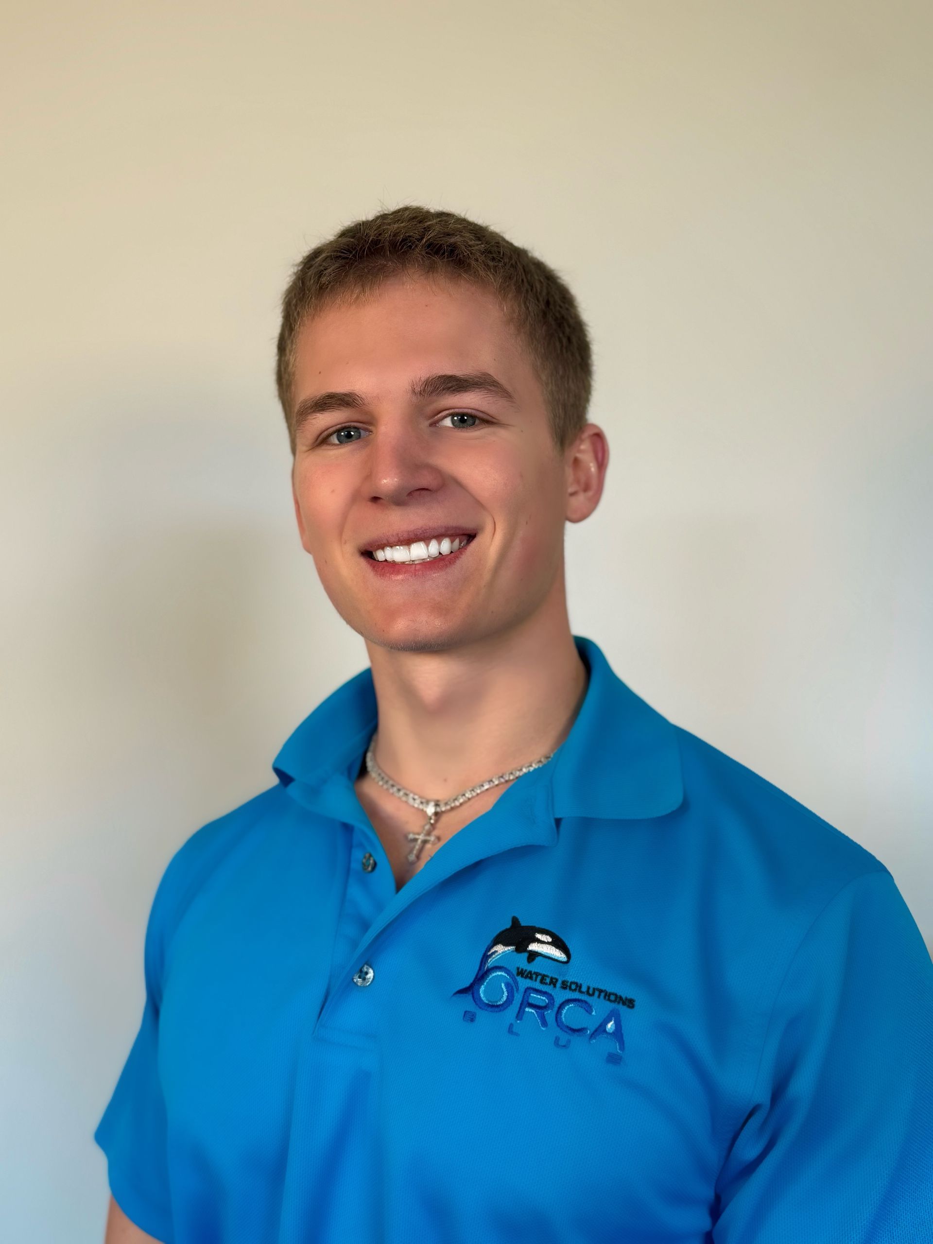 Smiling person in a blue polo shirt with a logo, standing against a plain beige background