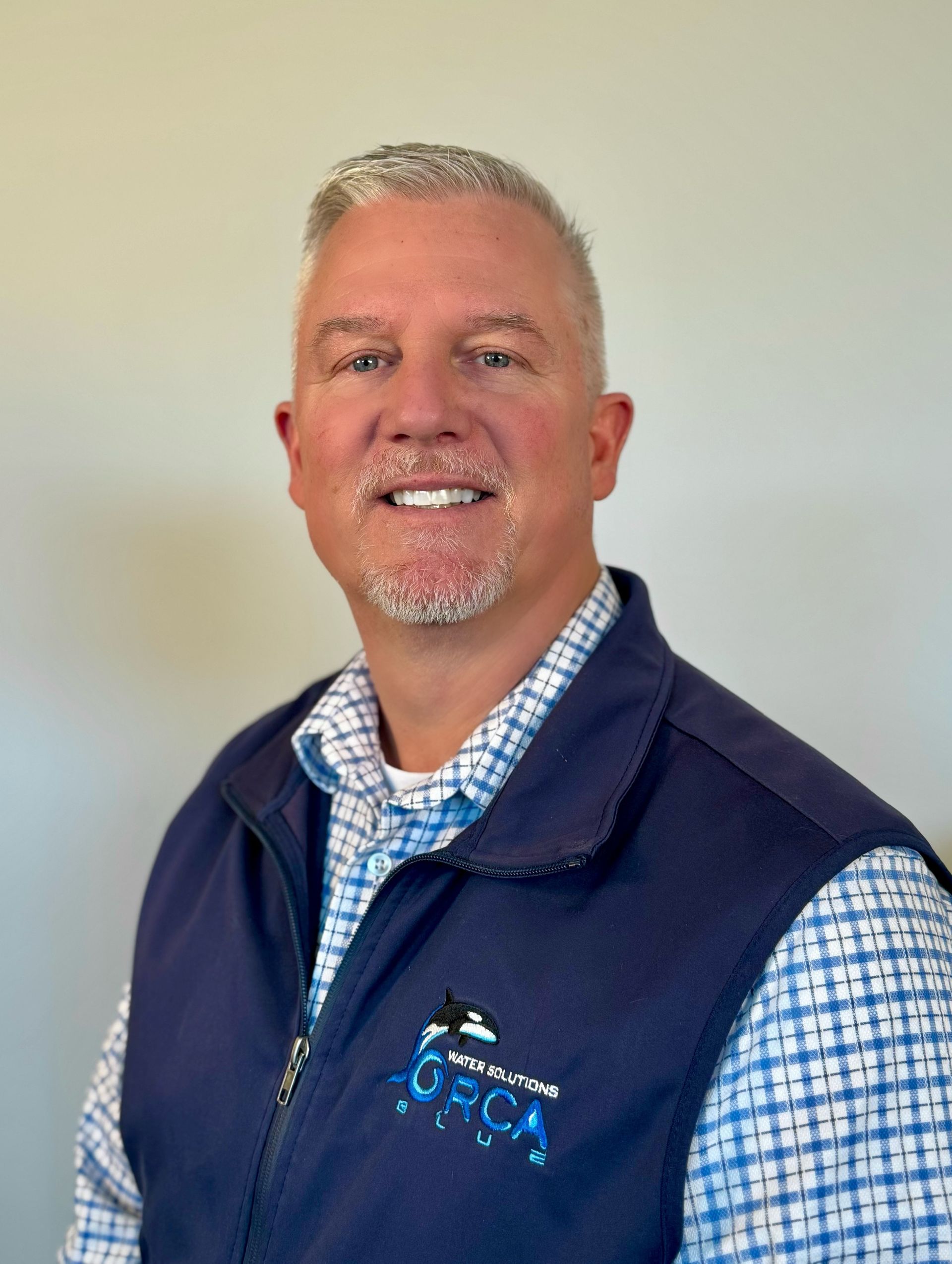 Smiling man in a navy vest and blue checkered shirt against a plain light wall