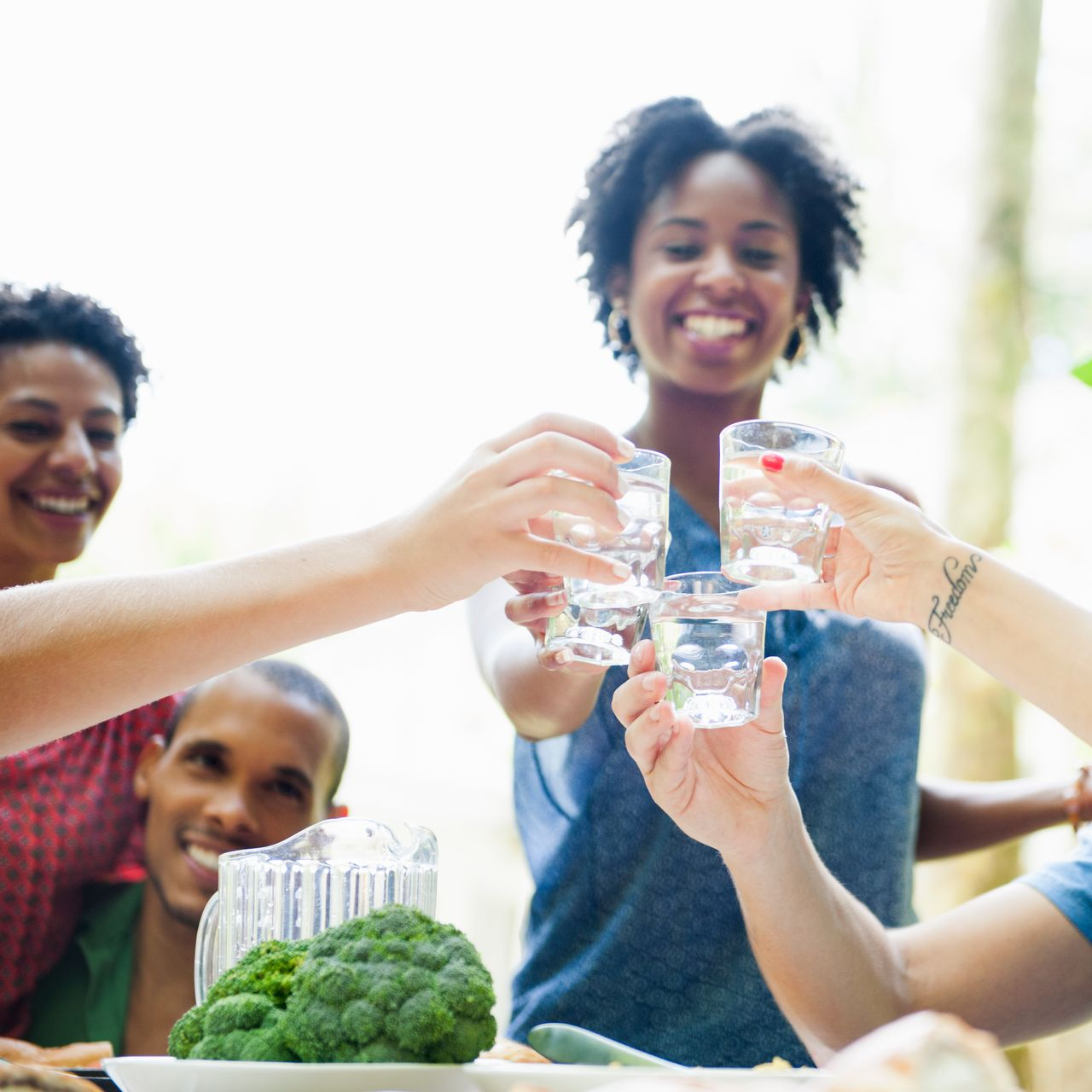 Friends clinking glasses of water at a bright outdoor picnic table with broccoli in the foreground