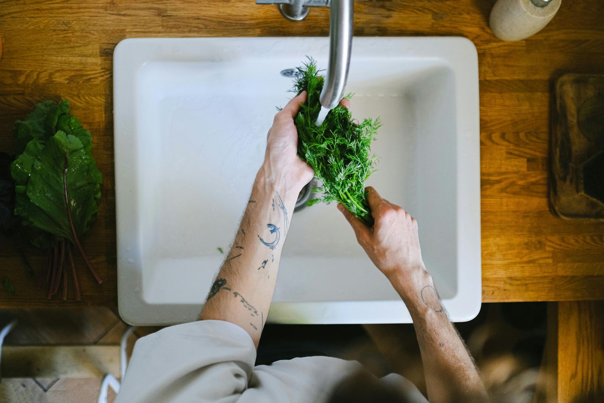 Hands washing leafy greens in a white kitchen sink under running water.
