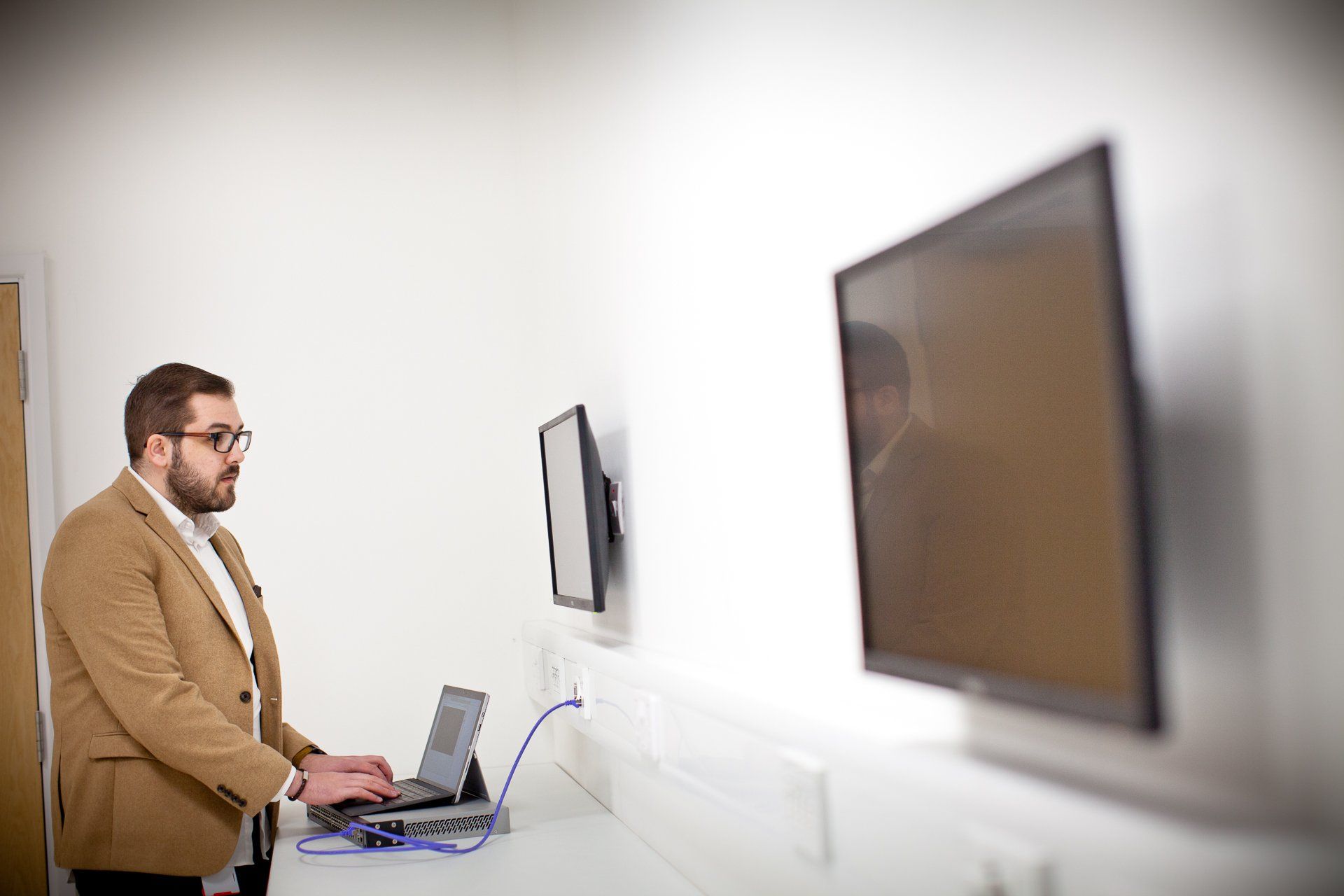 Man in blazer using laptop, facing two wall-mounted screens in a white room.