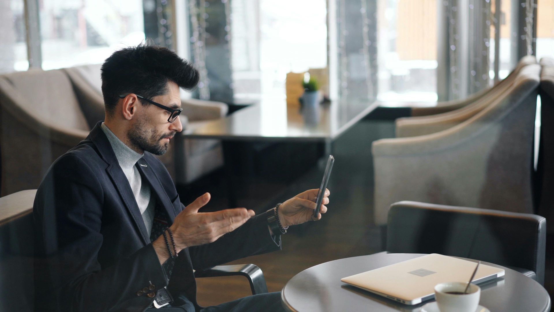 Man in suit on video call, gesturing with hand, sitting in a cafe with laptop and coffee.