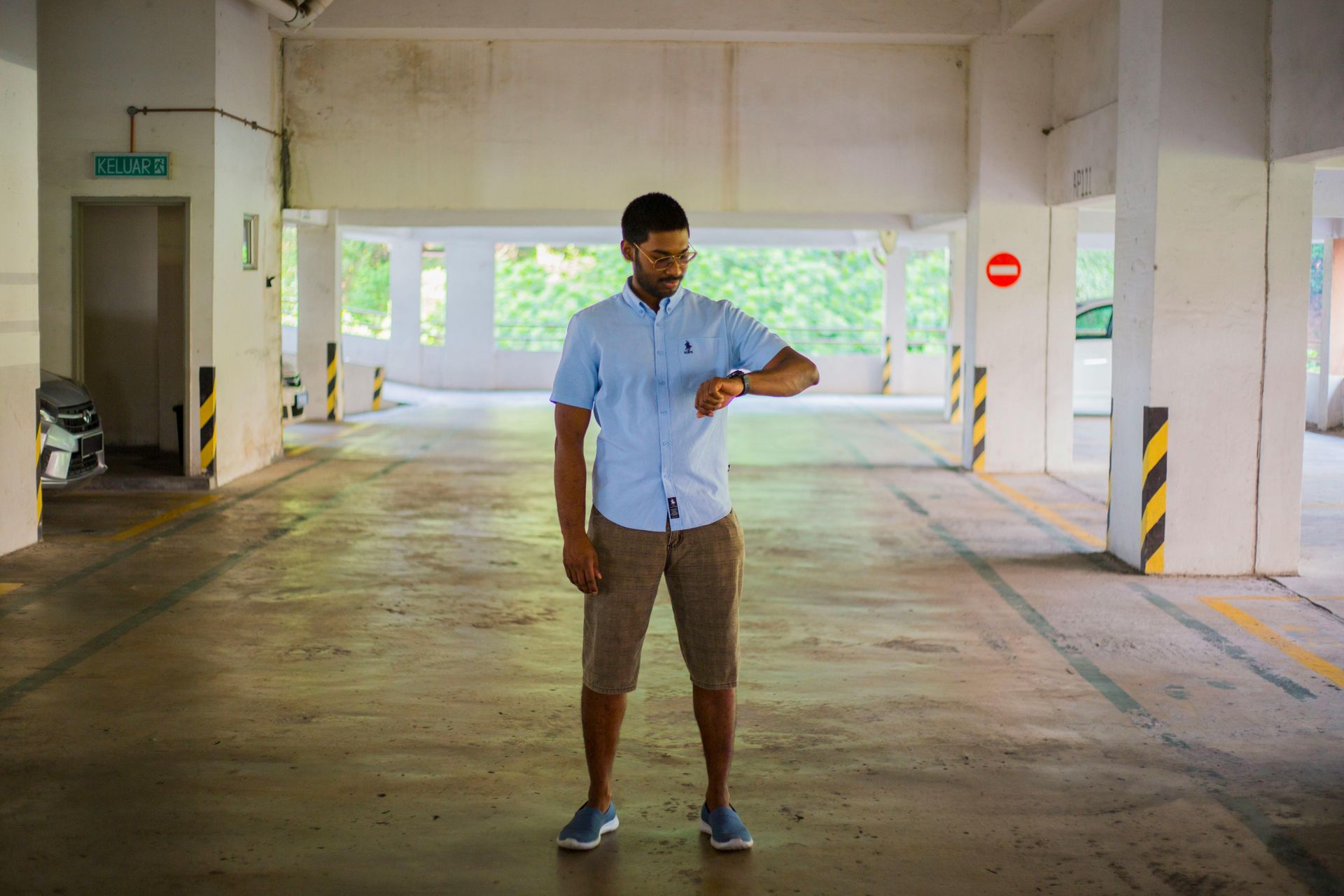 Man in blue shirt and shorts checks watch in a parking garage.