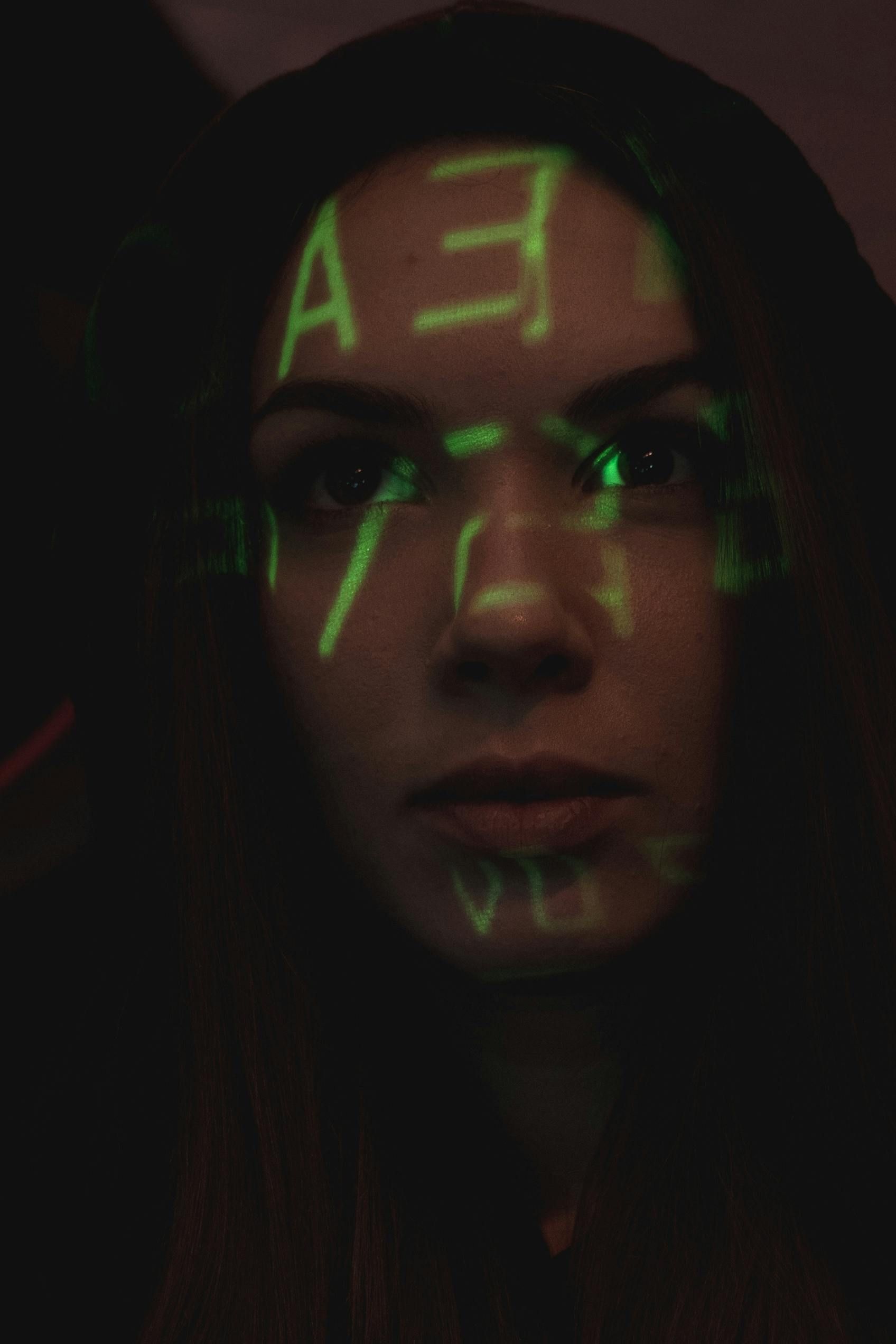 Woman with hooded sweatshirt, illuminated by green code projected on her face. Serious expression, dark background.