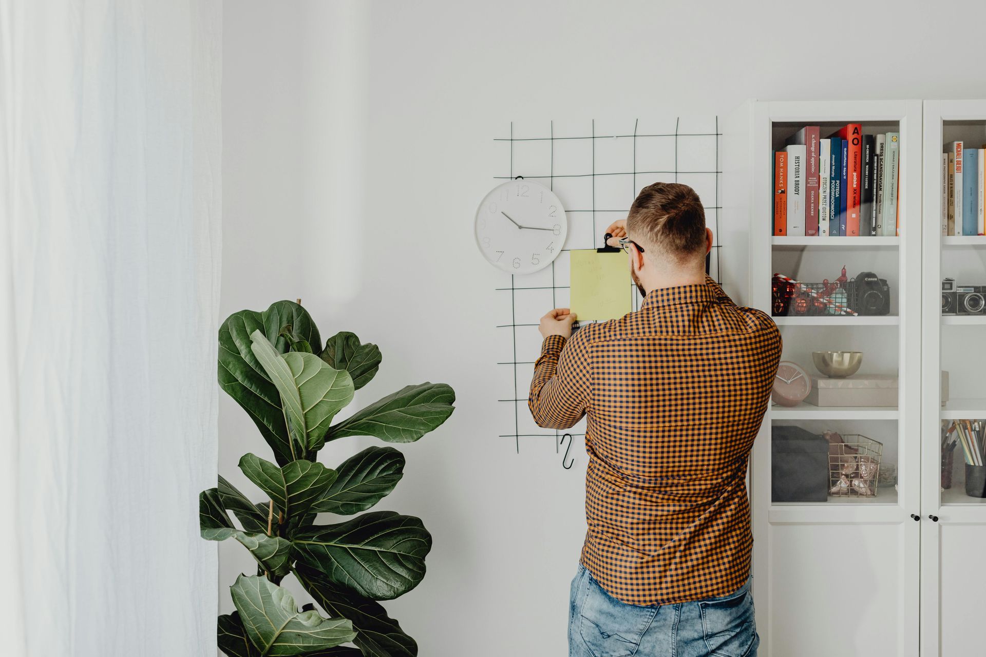 Man in orange plaid shirt hangs paper on wall near a bookcase and large plant.