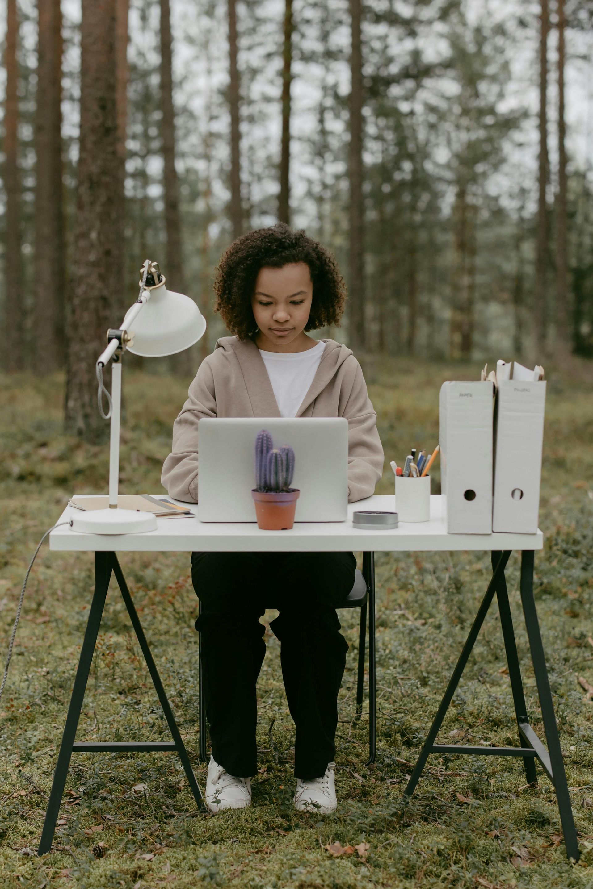 Person working on a laptop at a desk in a forest setting. A lamp and cactus are on the desk.