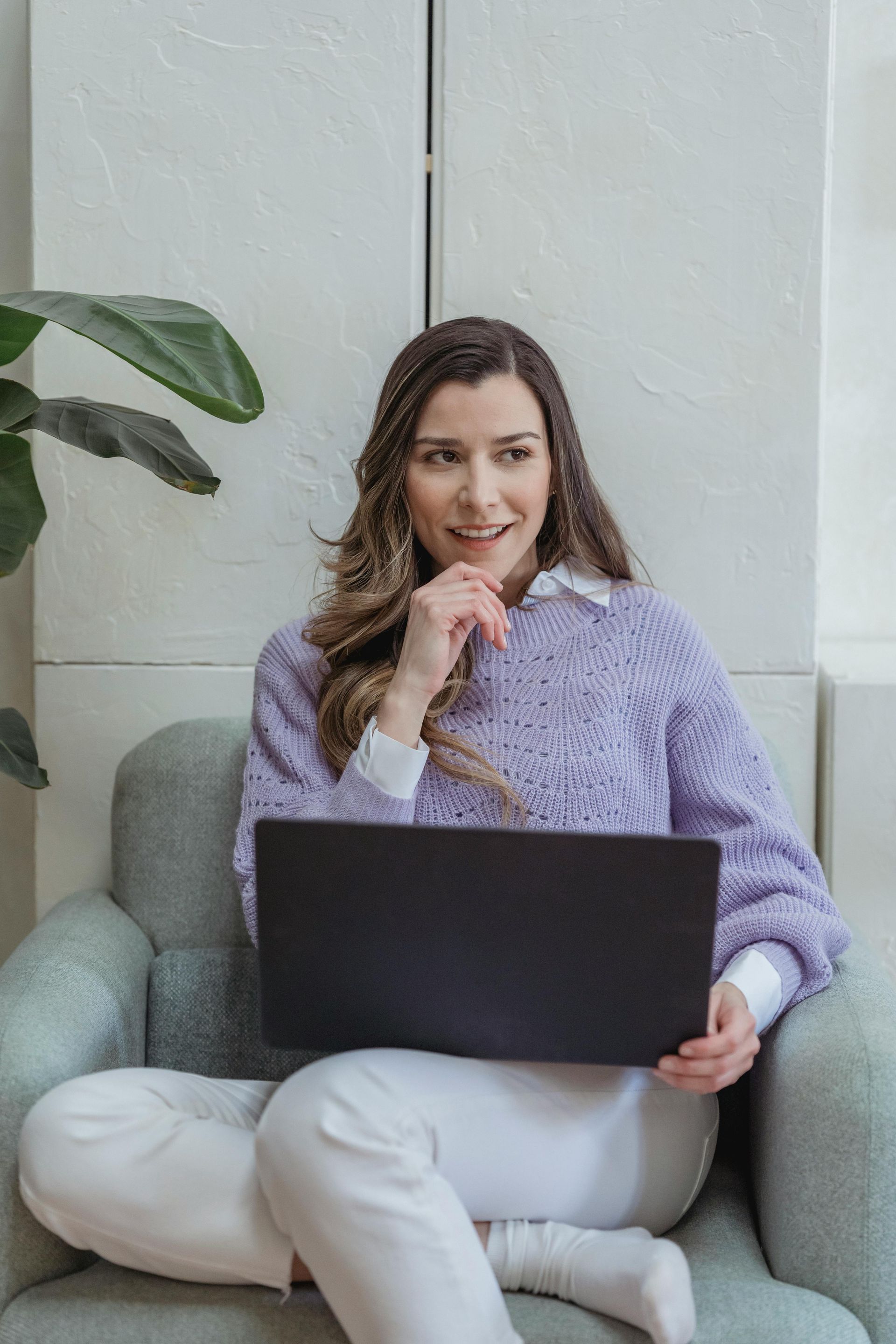 Woman in purple sweater, seated in armchair, using a laptop, looking thoughtful.