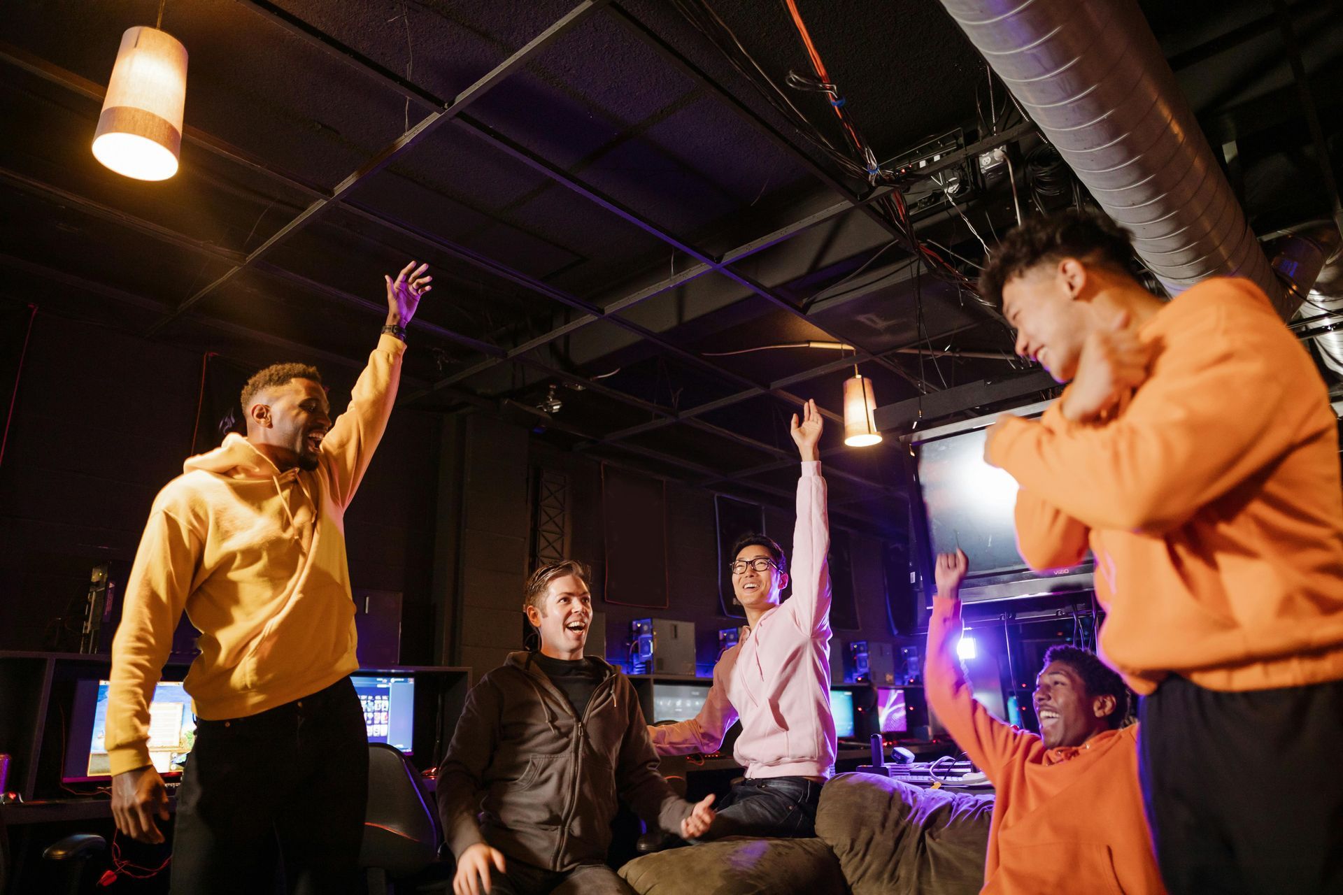 People celebrating in a room with computers, some with arms raised. Dim lighting, yellow and pink hoodies.