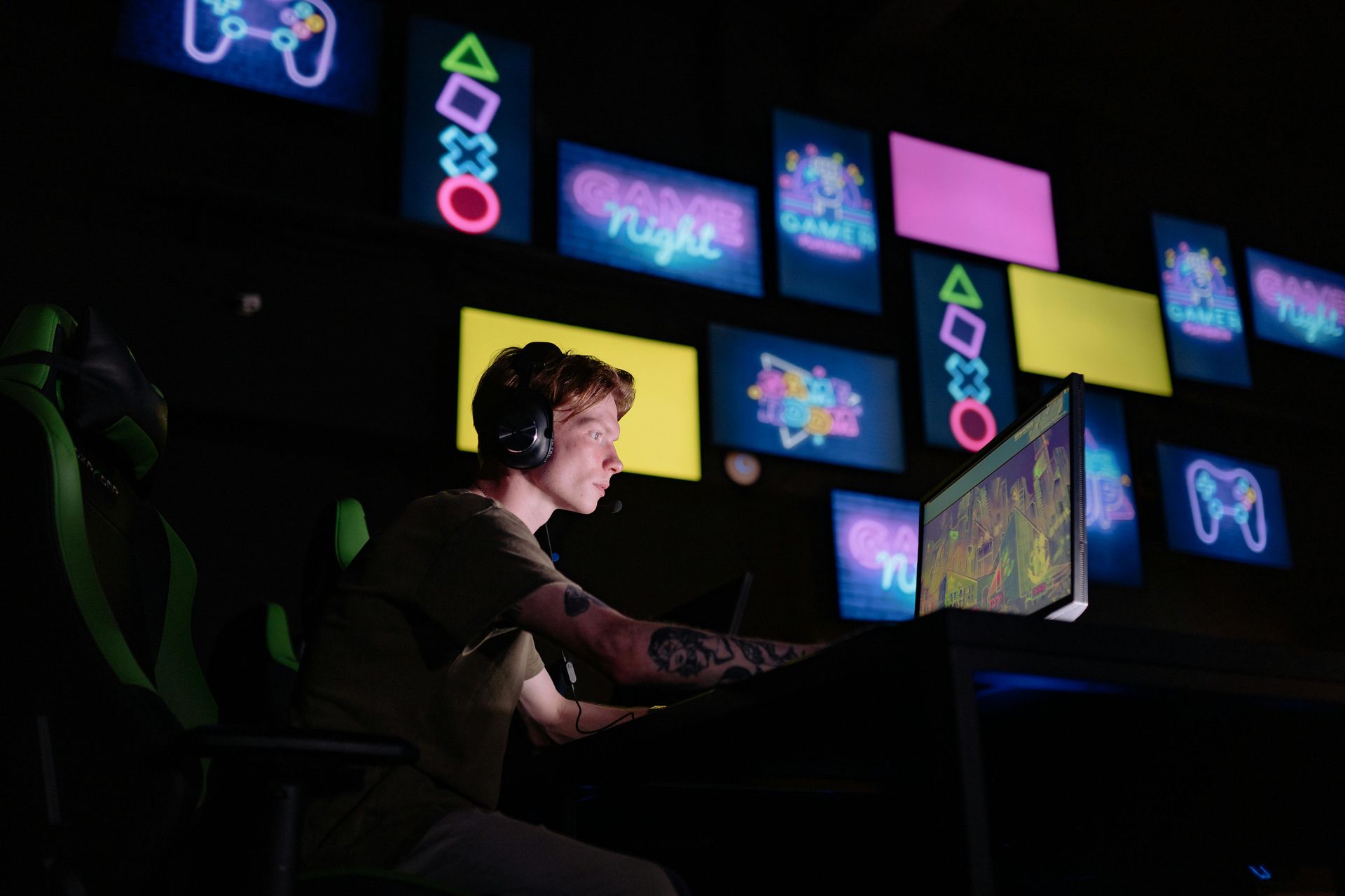 Person with headset, playing video game, illuminated by neon game art on dark wall.
