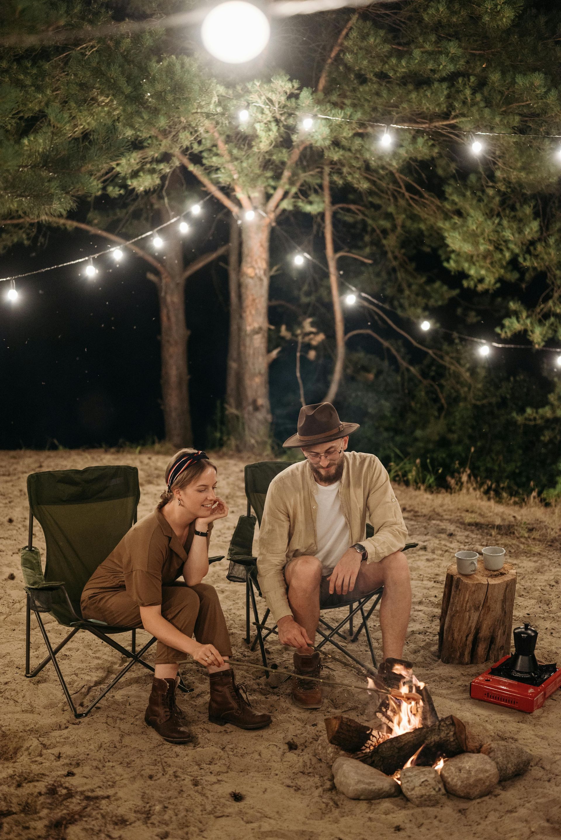 Couple camping near a fire pit, seated on chairs. String lights above.