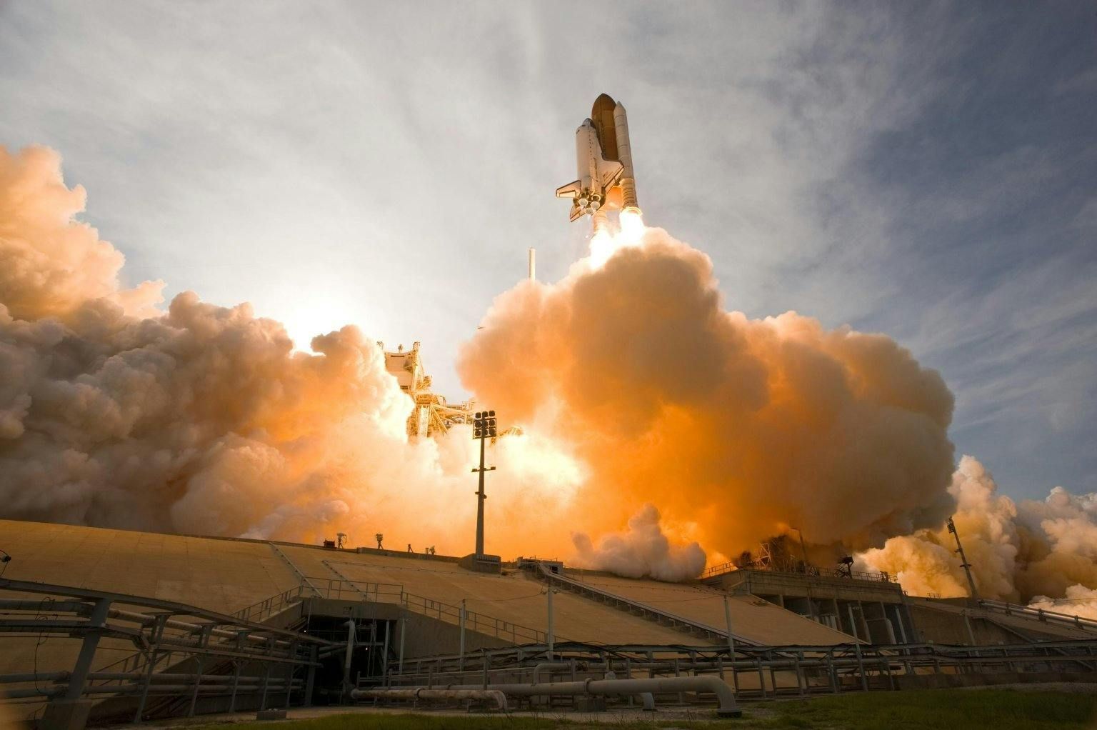 Space shuttle launching, engulfed in orange and white flames and smoke, rising above launchpad structures.