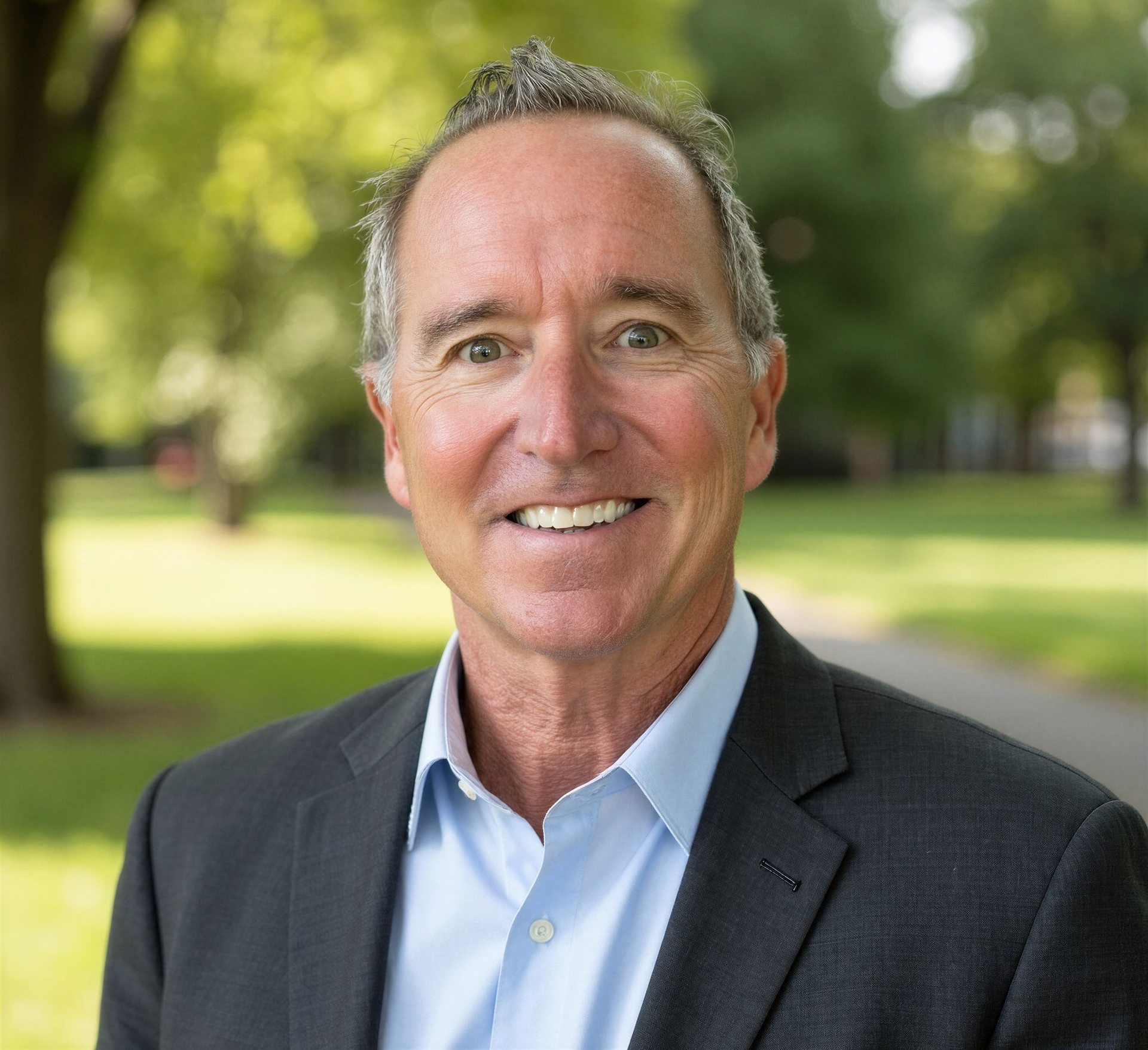Smiling man in a dark blazer and light blue shirt standing outdoors in a park