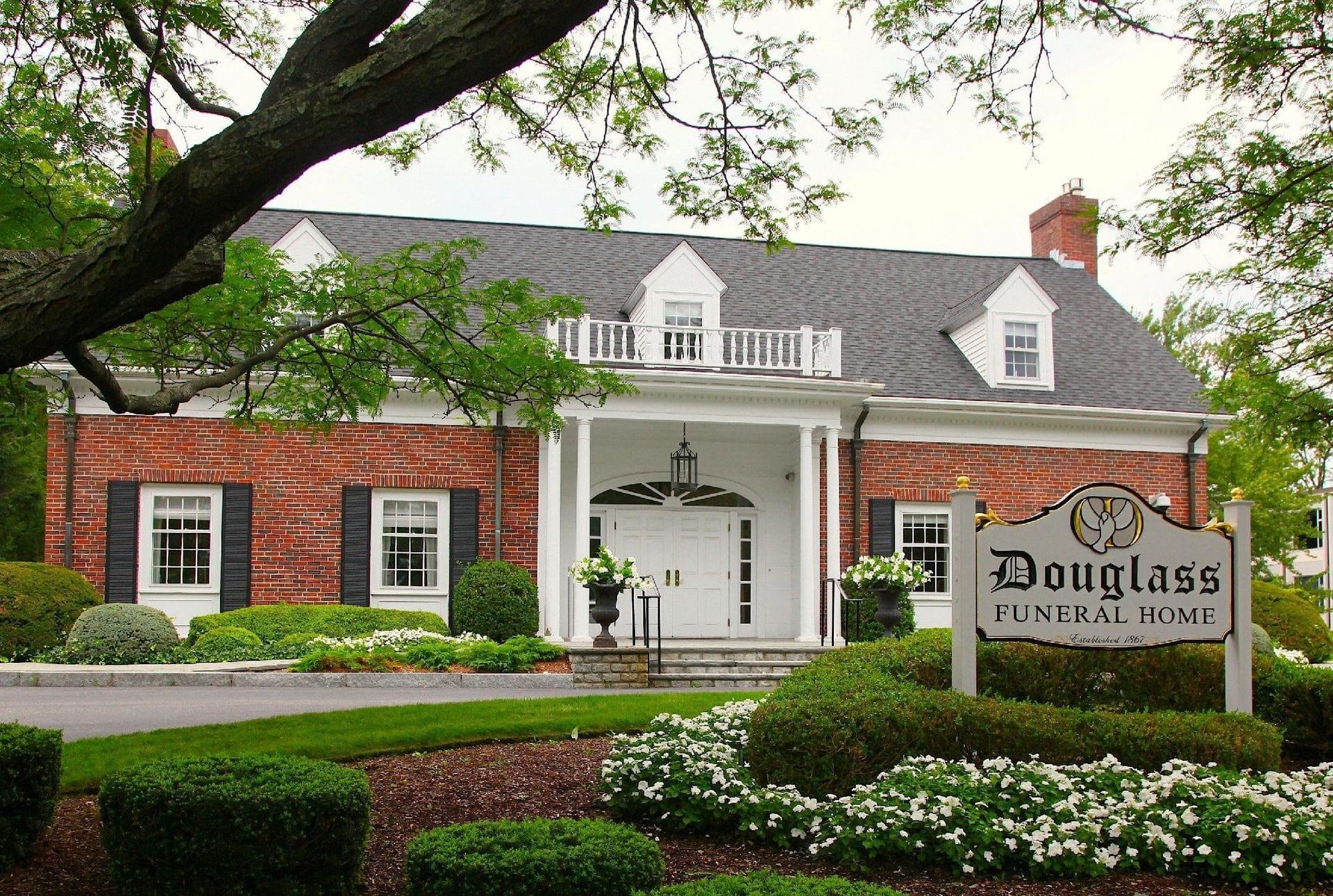 Douglass home-style building with white columns and sign, surrounded by trees and flowers