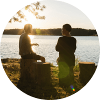 Two people sit by a lake at sunset, silhouetted against the water and trees.