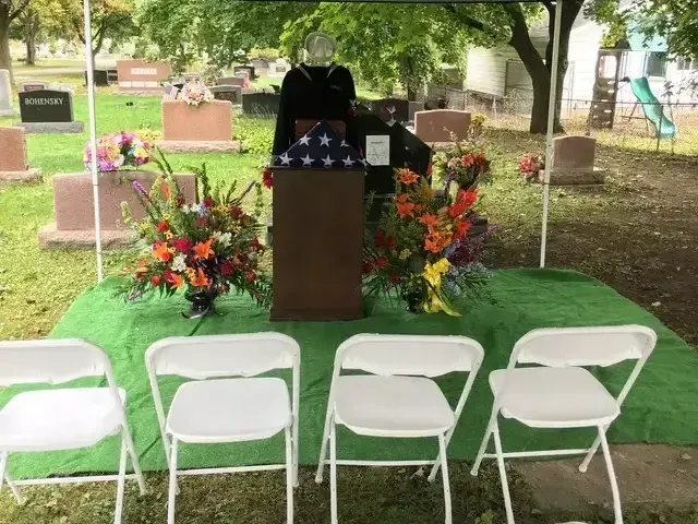 Memorial podium under trees with floral arrangements and four white folding chairs in front