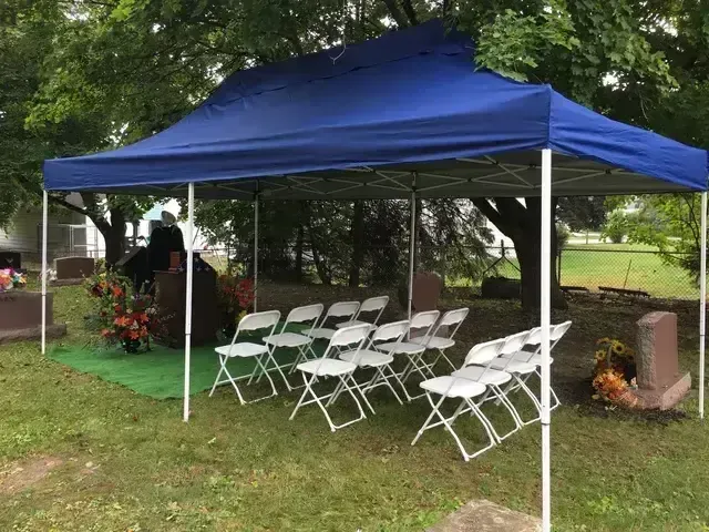 Blue pop-up canopy over white folding chairs in a grassy yard, with flowers and trees nearby.