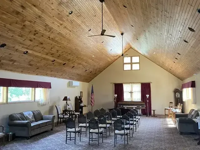 Spacious meeting hall with rows of chairs, a piano, flags, and sofas under a wood-paneled vaulted ceiling