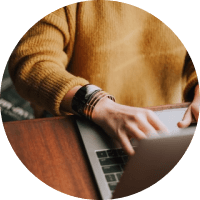 Person in a mustard sweater typing on a laptop at a wooden table.