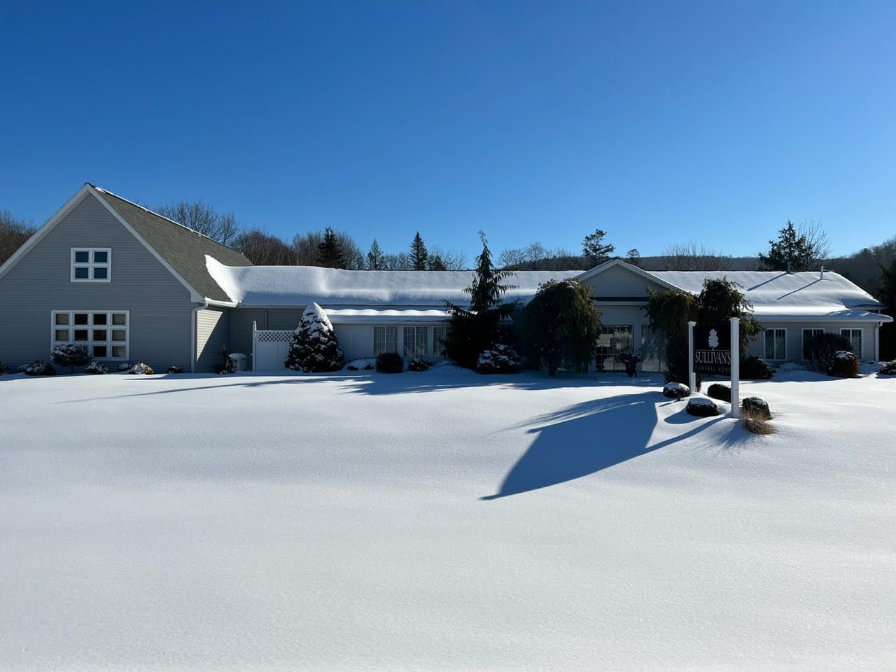 Snow-covered suburban house under a clear blue sky, with a long driveway and dark shutters