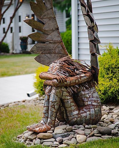 Winged metal garden sculpture on a rock bed beside a house and lawn