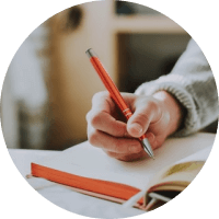 Hand holding a red pencil over paper beside an open notebook