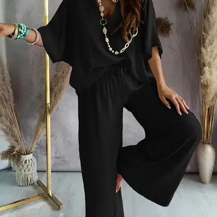 Woman in a white loose blouse and wide-leg pants standing in a bright room with mirrors and dried grasses
