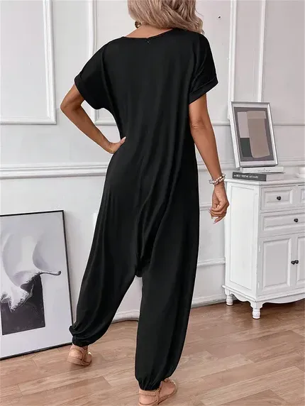 Person in a black loose jumpsuit standing in a bright room with a dresser and framed art.