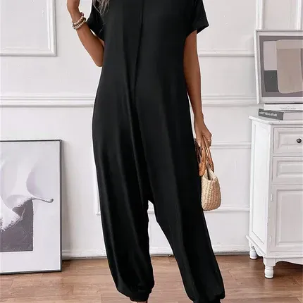 Person in a black loose jumpsuit standing in a bright room with a dresser and framed art.