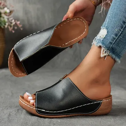 Black and tan slip-on sandals, one held in hand and one worn on a foot in ripped jeans
