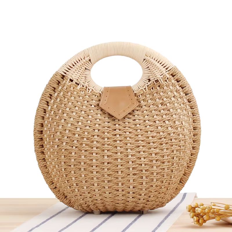 Round woven straw handbag with a tan leather accent, standing on a light wooden surface.