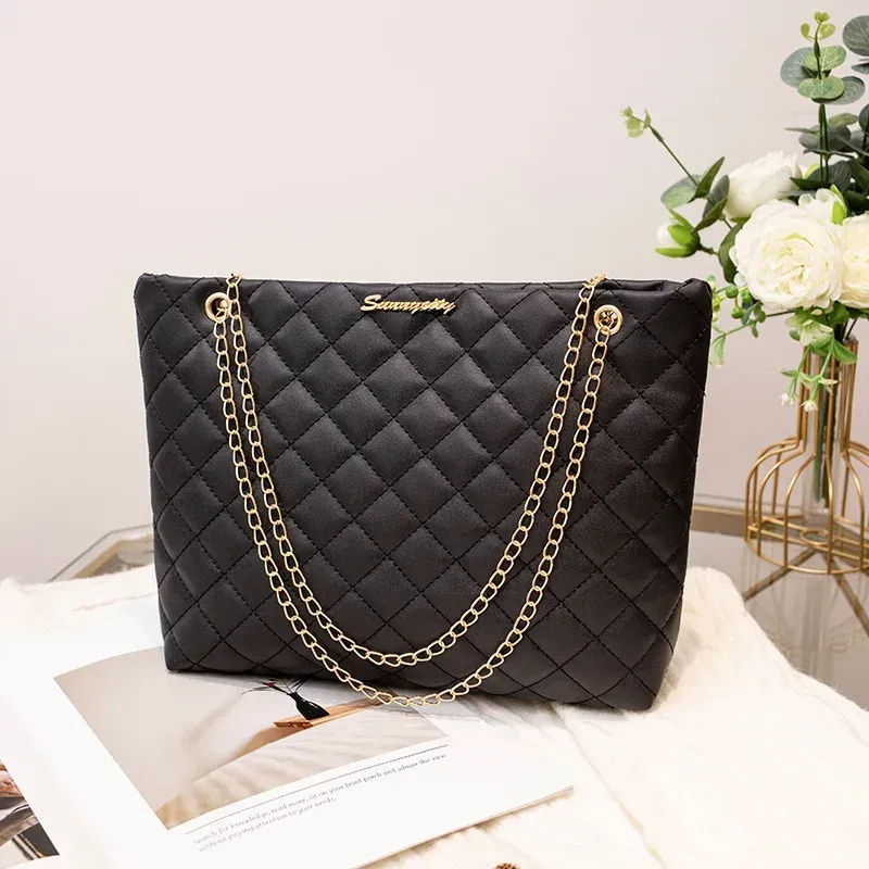 Black quilted handbag with gold chain straps on a white surface beside flowers and a magazine