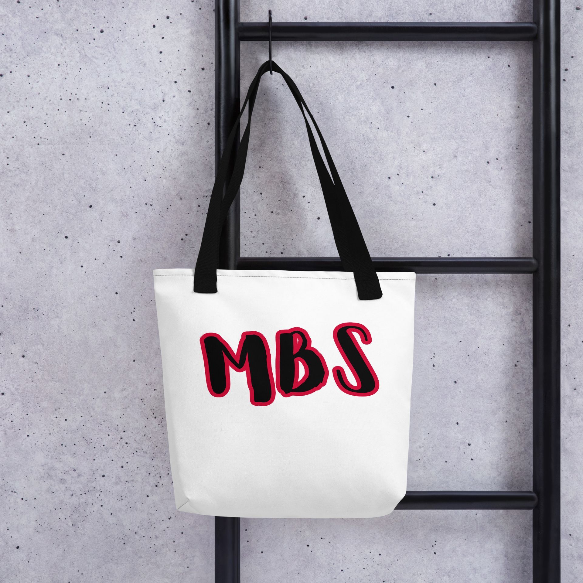 White tote bag with black handles and red “MBS” lettering on a plain background