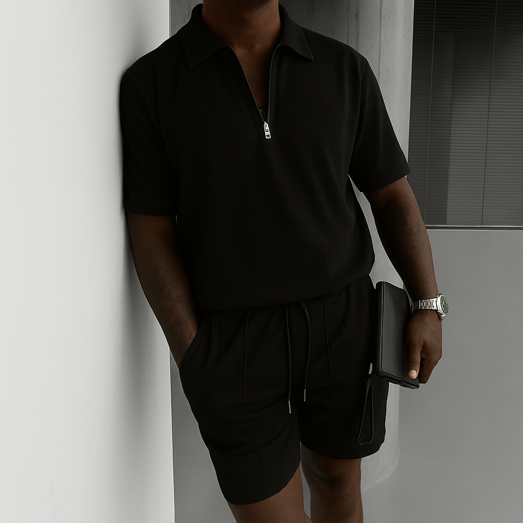 Person in a black polo and shorts leaning against a wall in a minimalist indoor setting