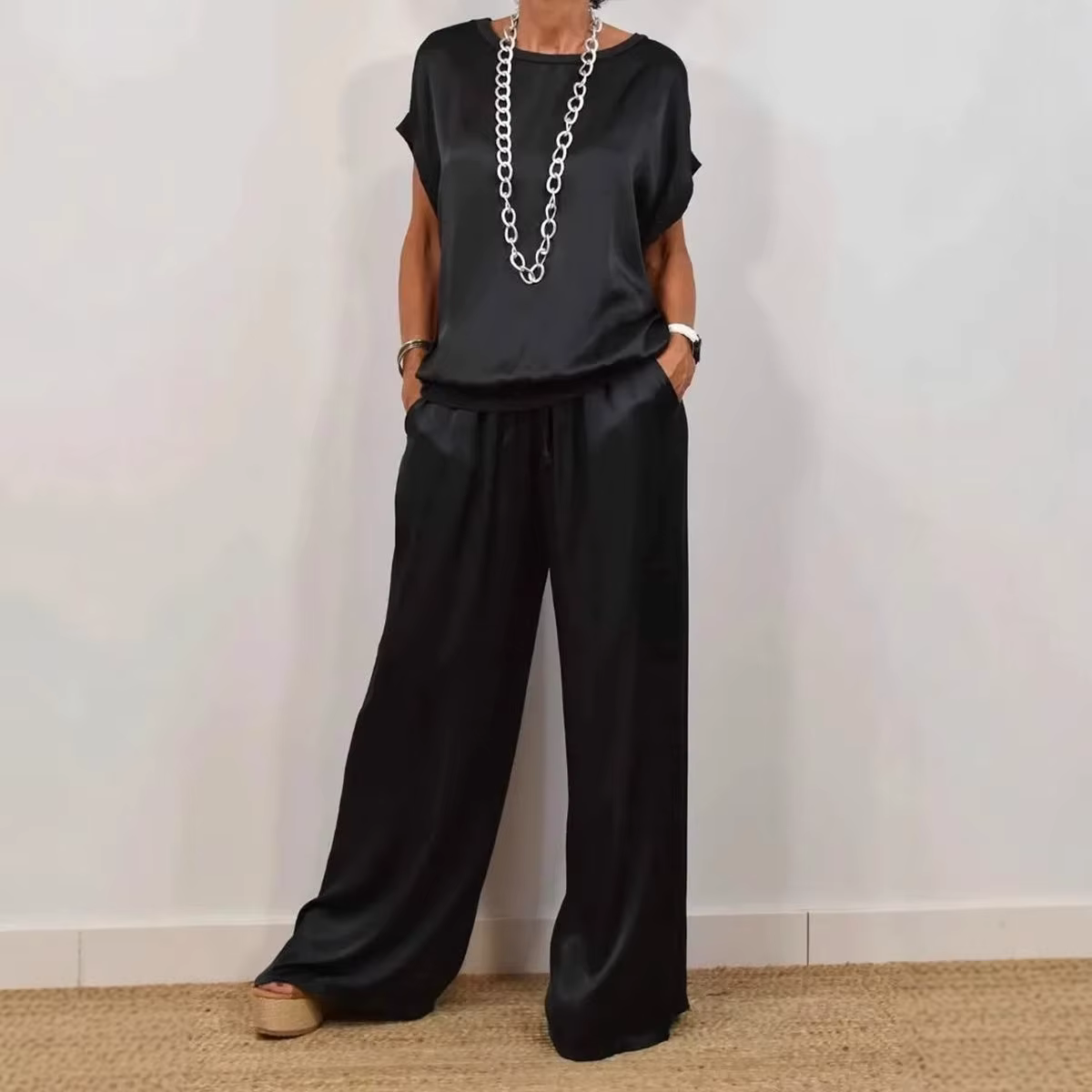 Person in a black loose-fit outfit standing against a light wall, hands in pockets, wearing a long silver necklace.