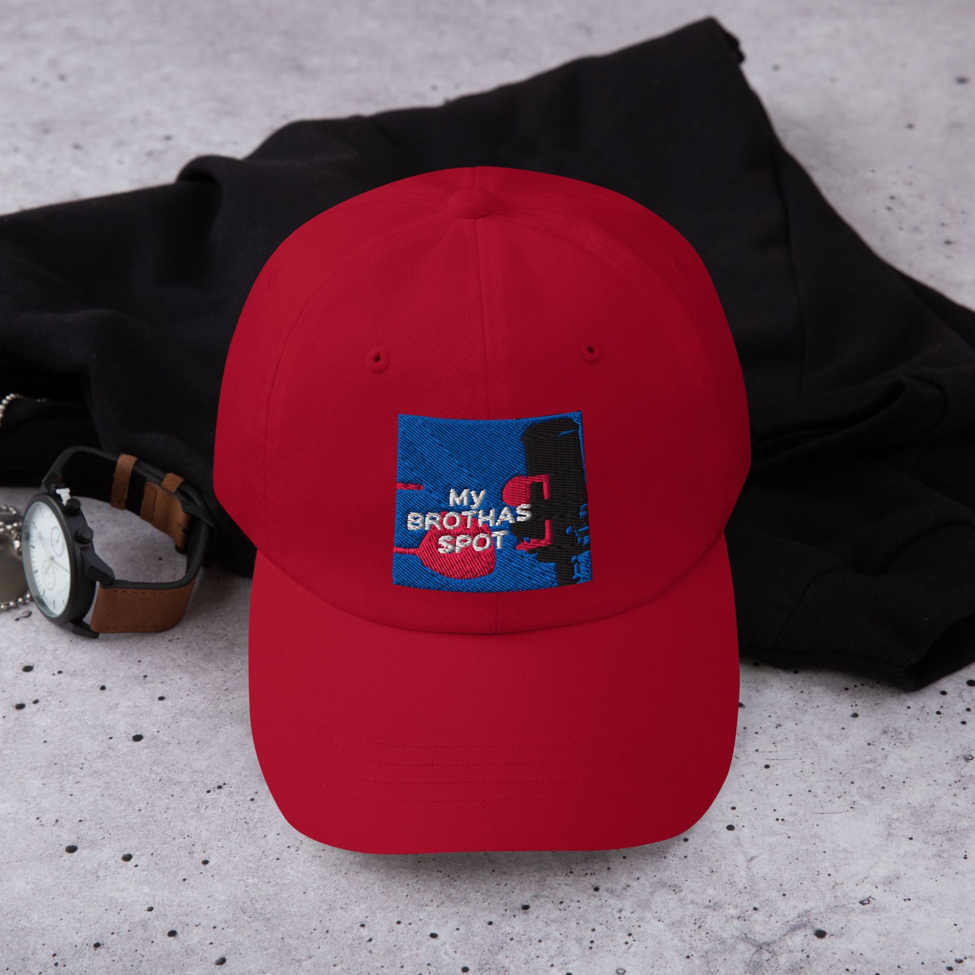 Red baseball cap with a colorful patch, resting on a dark cloth beside sunglasses.