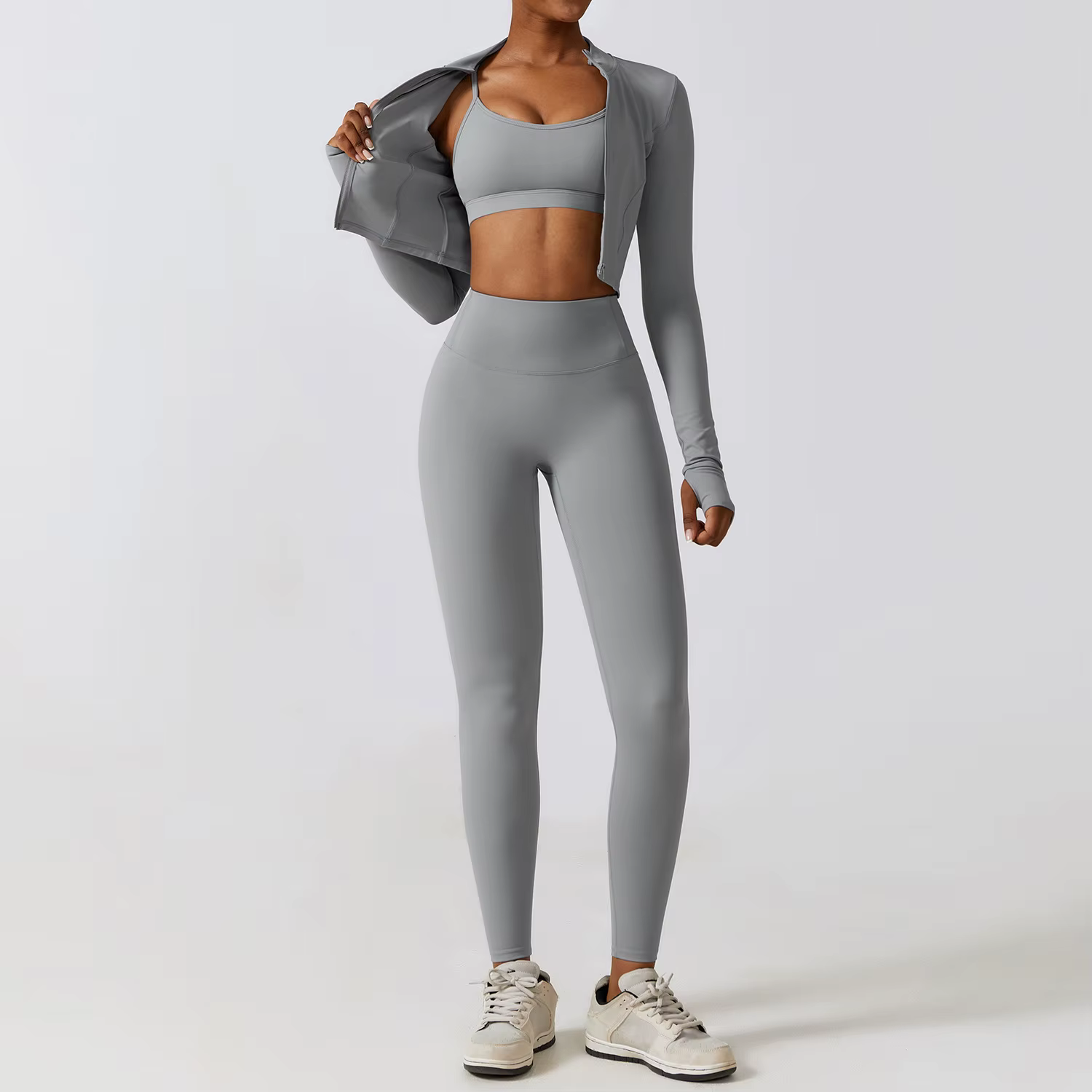 Person in gray athletic wear standing in a studio, holding a jacket open, wearing sneakers.