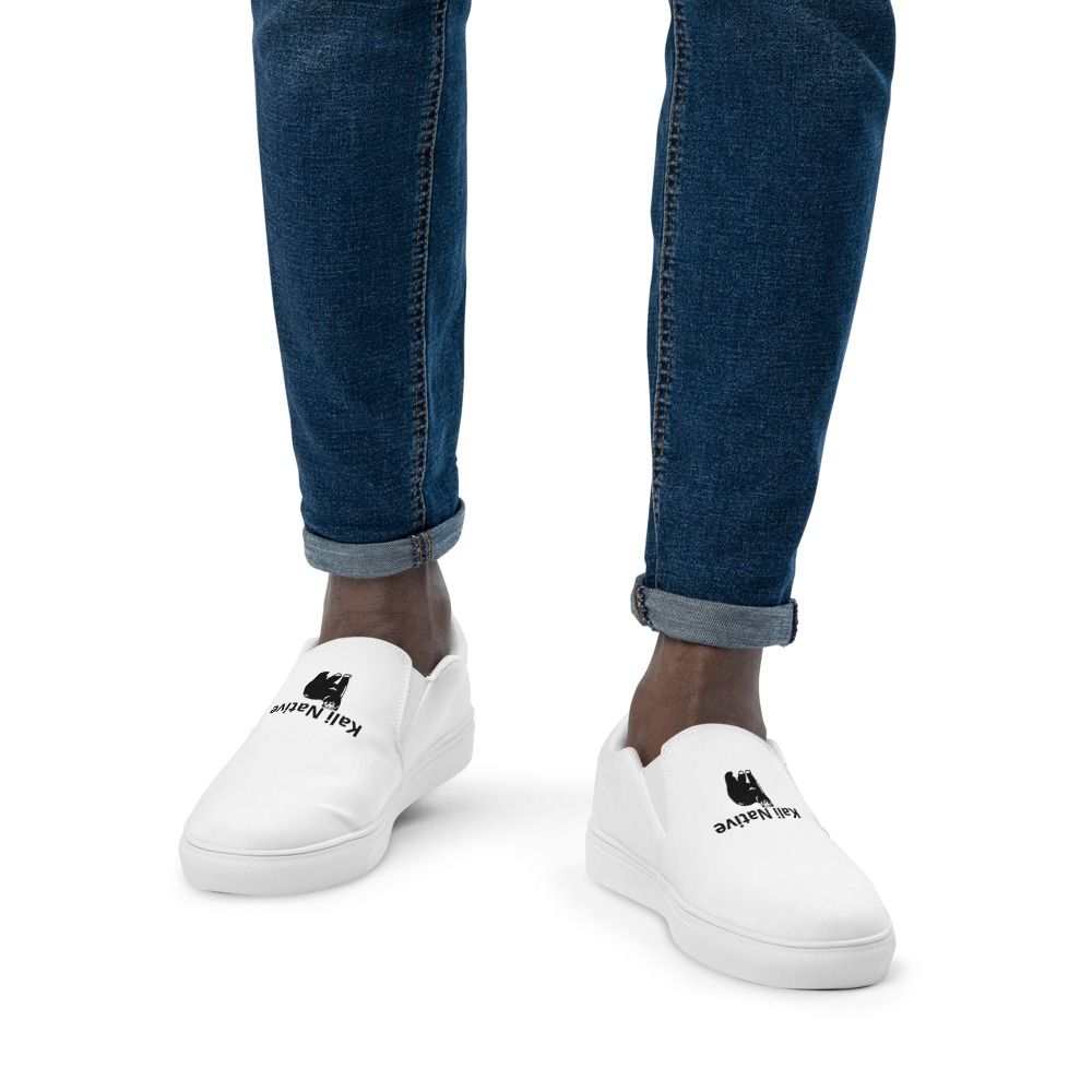 White slip-on sneakers with black logos, worn with rolled-up blue jeans on a white background