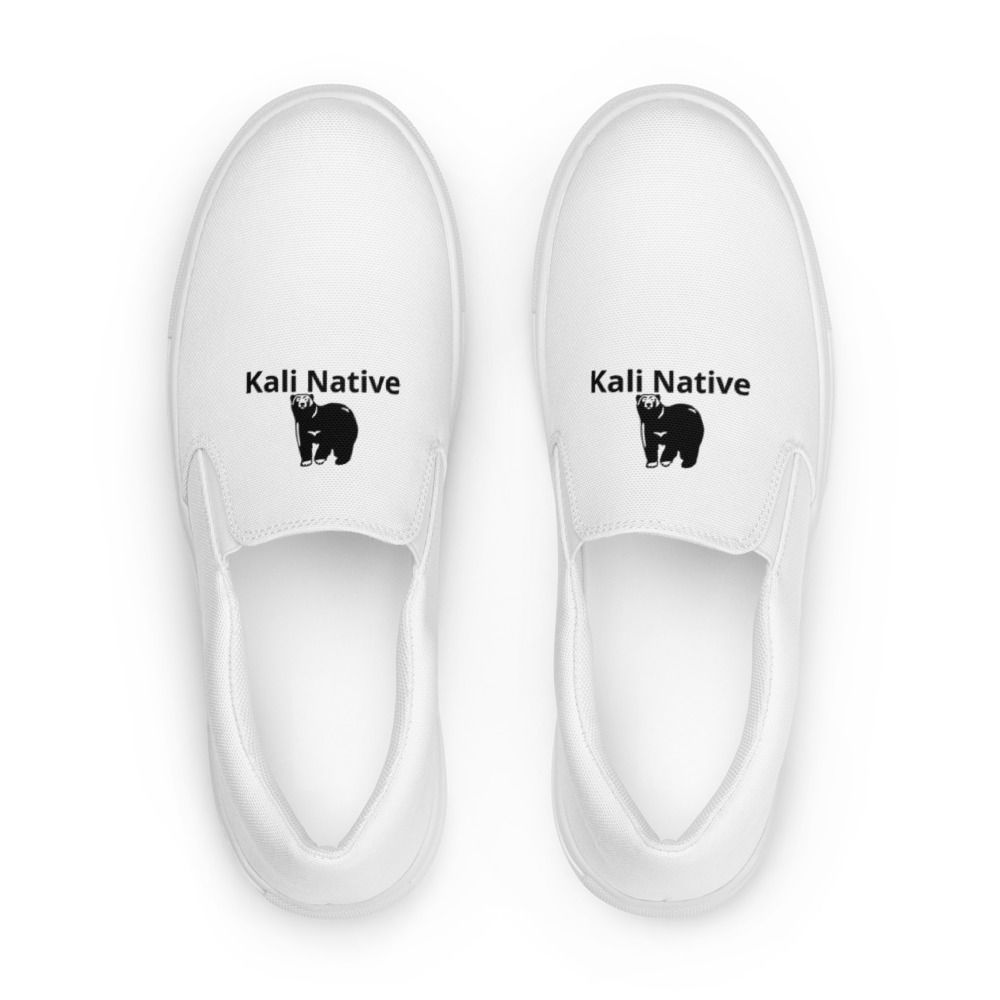 White slip-on sneakers with black logos, worn with rolled-up blue jeans on a white background